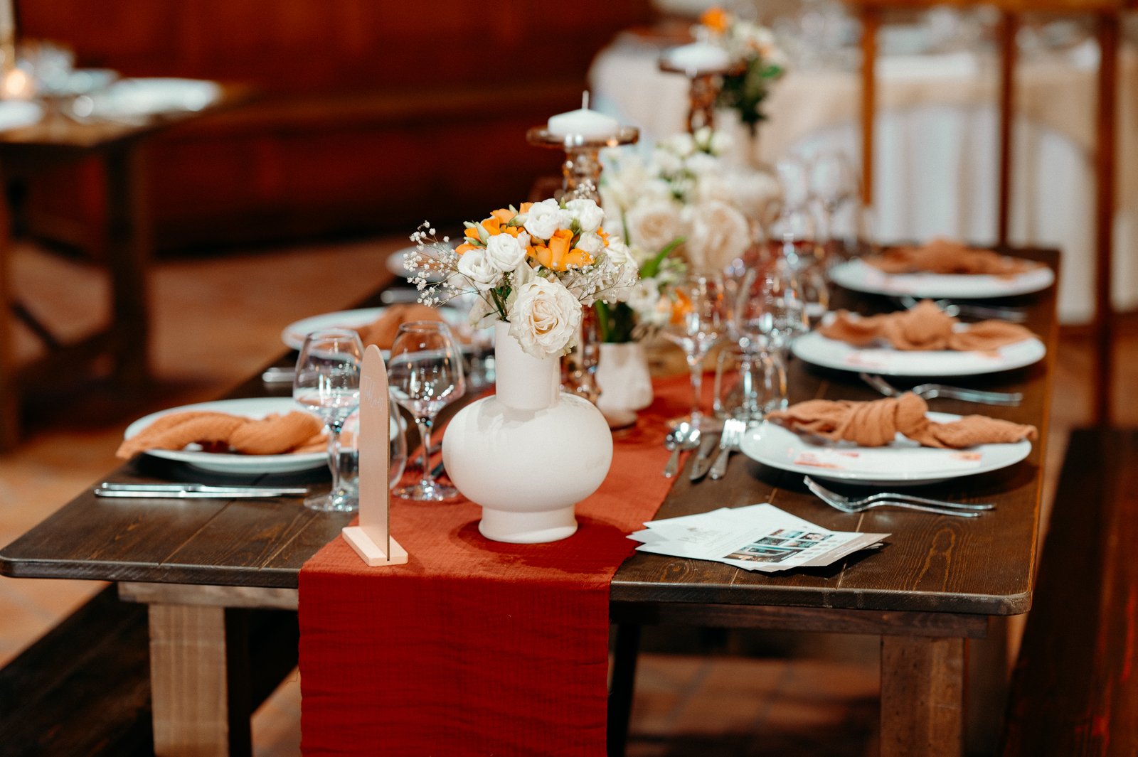 Vase boule en céramique blanche garni de roses blanches et orange sur un chemin de table terracotta, table en bois