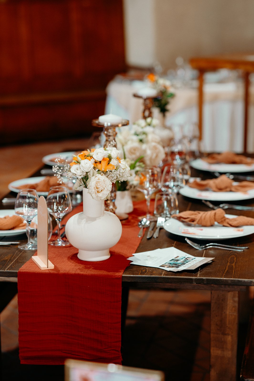 Tablée en bois dressée d’assiettes blanches et de serviettes caramel, vase blanc et chemin de table terracotta tombant