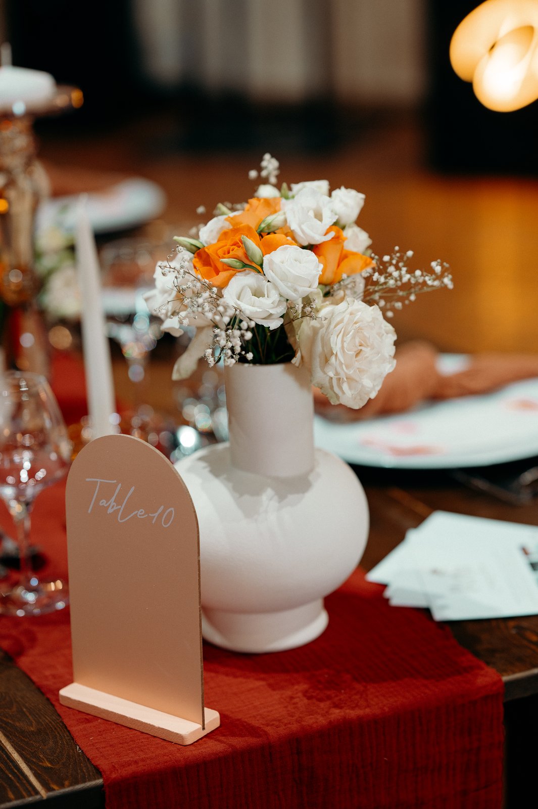 Numéro de table en arche couleur nude devant un vase blanc de roses orange, roses blanches et gypsophile