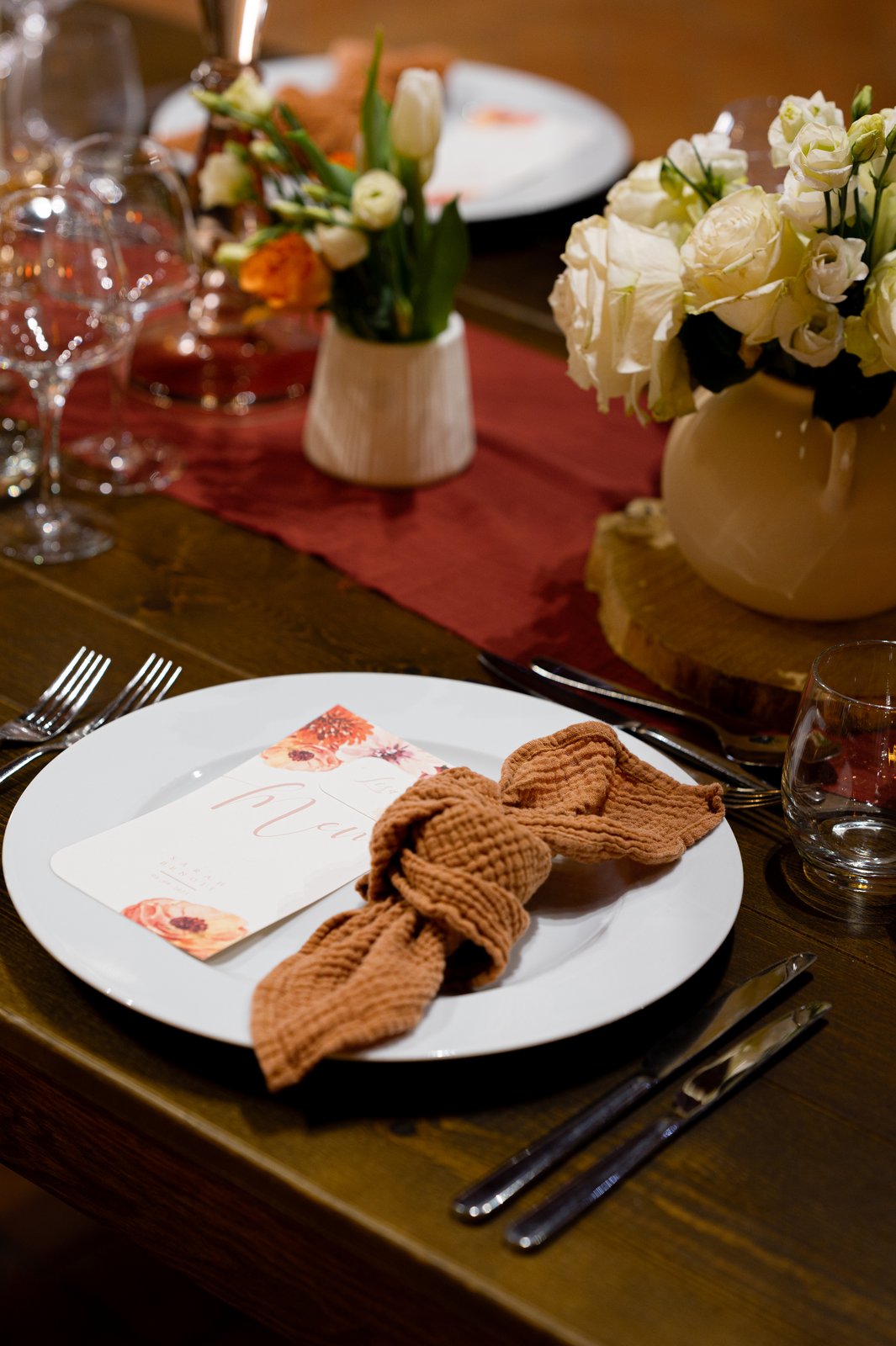 Assiette blanche, menu fleuri et serviette en gaze nouée, petits vases en céramique blanche et roses sur une table en bois