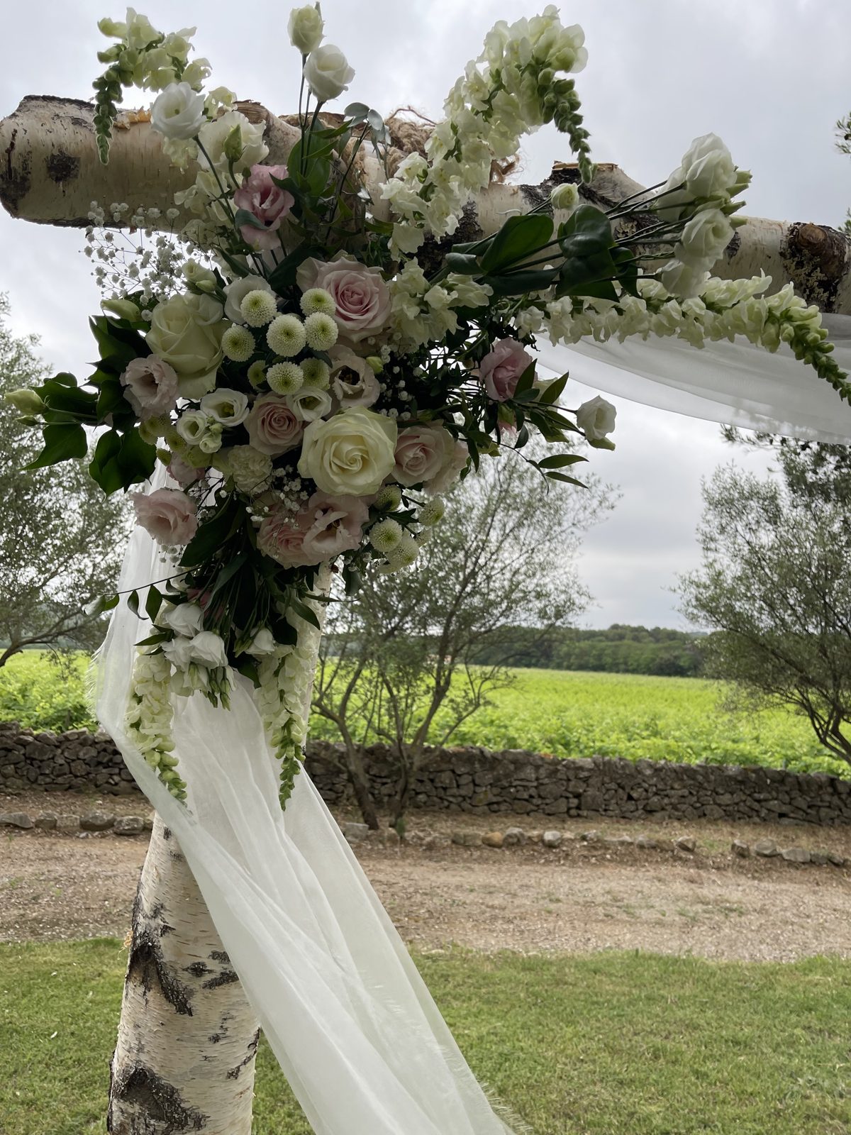 Composition de roses, lisianthus et gueules-de-loup blanches à l’angle d’une arche en bouleau drapée de voile