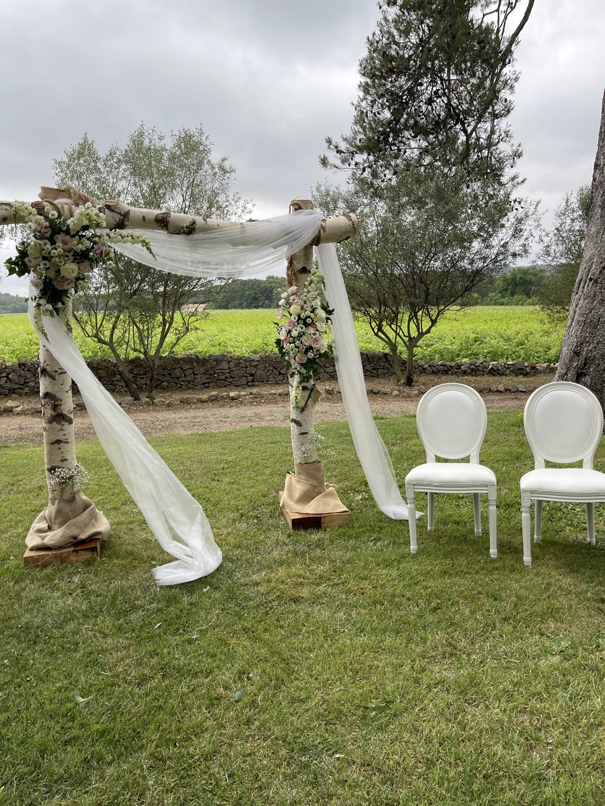 Arche en bouleau blanc drapée de voile et deux chaises médaillon blanches pour les mariés sur la pelouse du domaine