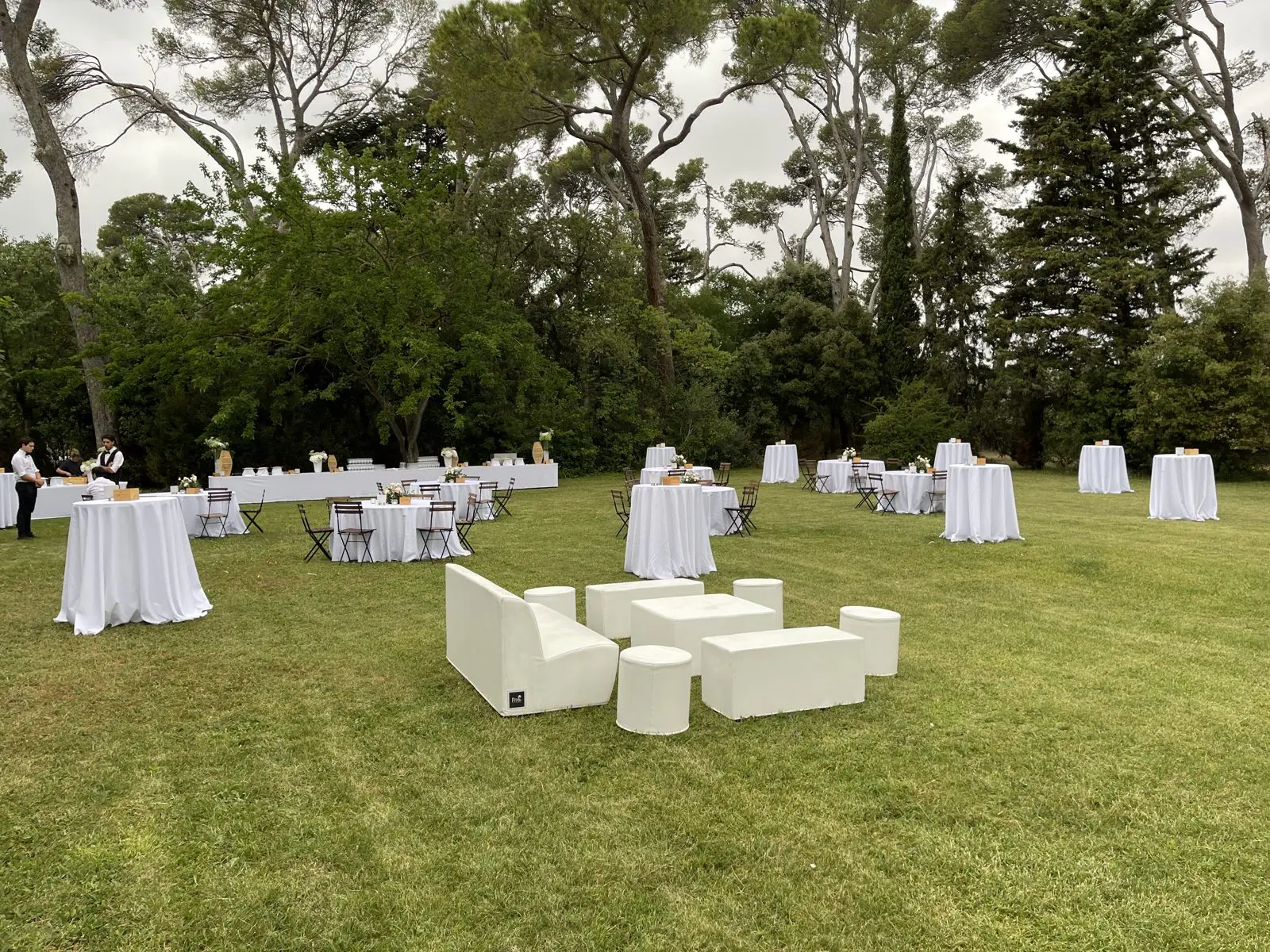 Salon lounge blanc et mange-debout nappés de blanc répartis sur la pelouse d’un parc arboré pour un cocktail