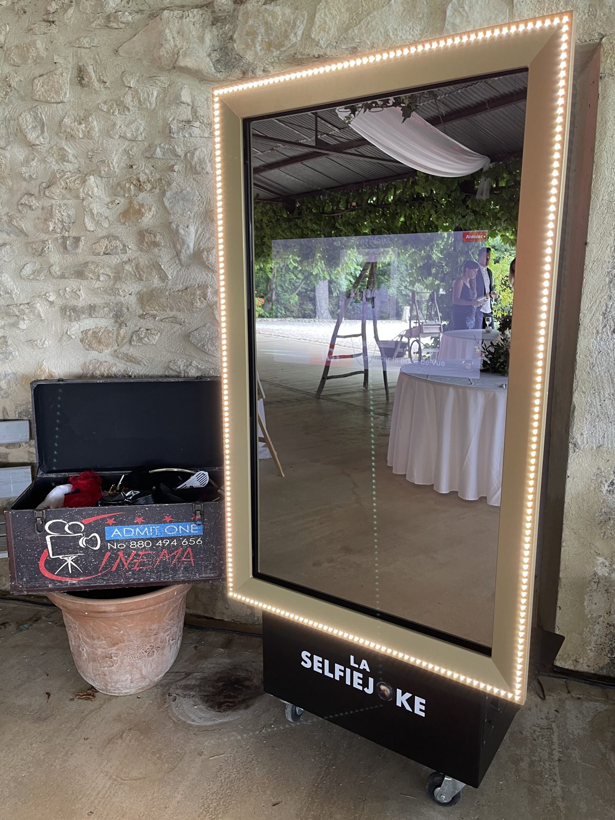 Borne photo miroir encadrée de lumières LED devant un mur de pierre, malle d’accessoires posée à côté