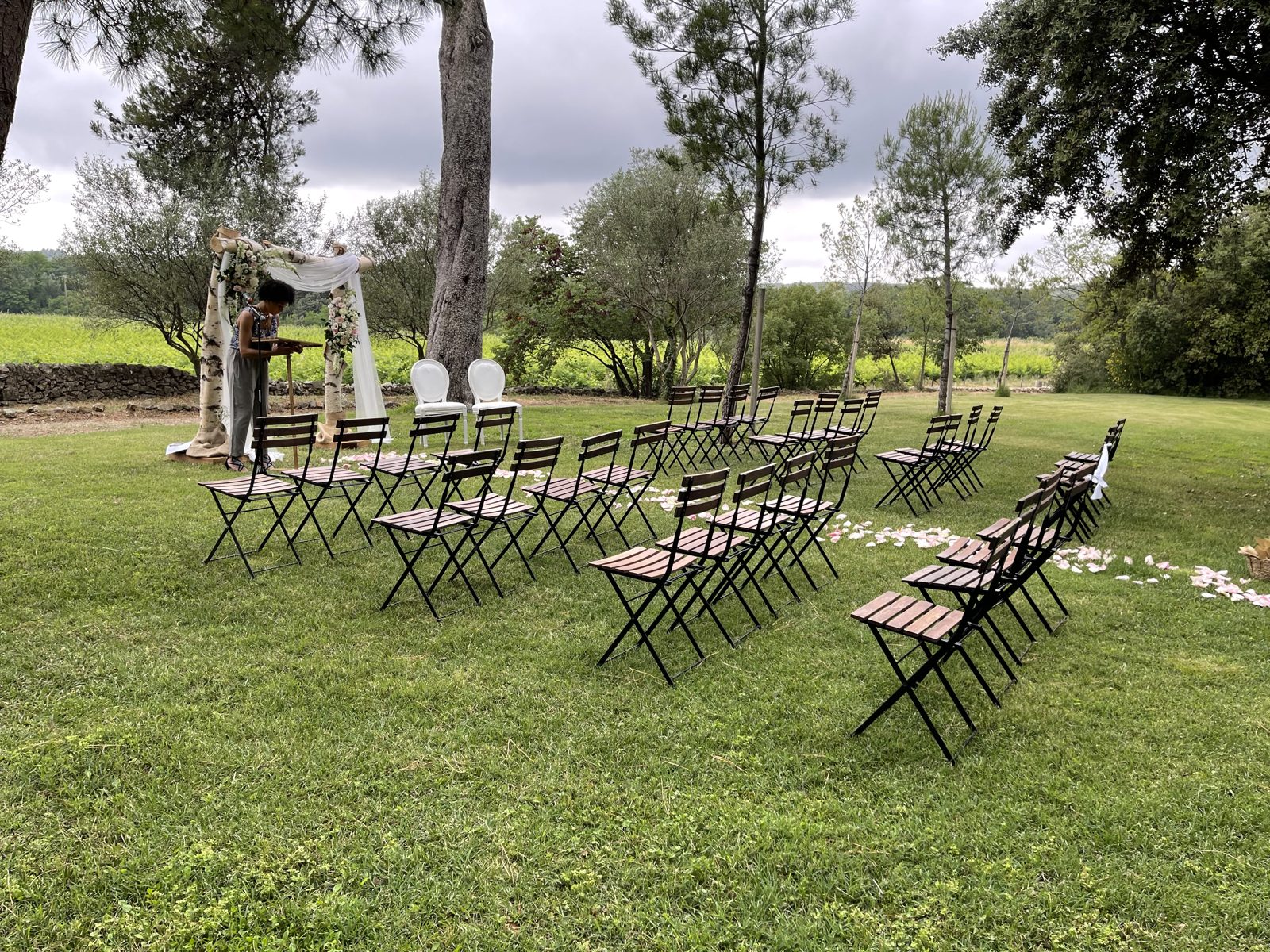 Rangées de chaises pliantes en bois face à une arche en bouleau fleurie, pupitre et pétales sur la pelouse du domaine