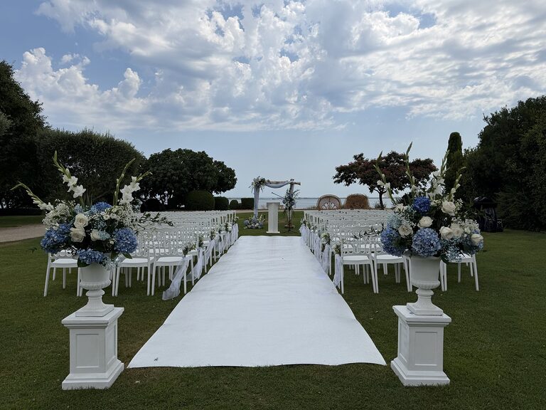 Cérémonie sur pelouse face à l’eau : allée blanche entre chaises blanches et vases Médicis d’hortensias bleus sur colonnes