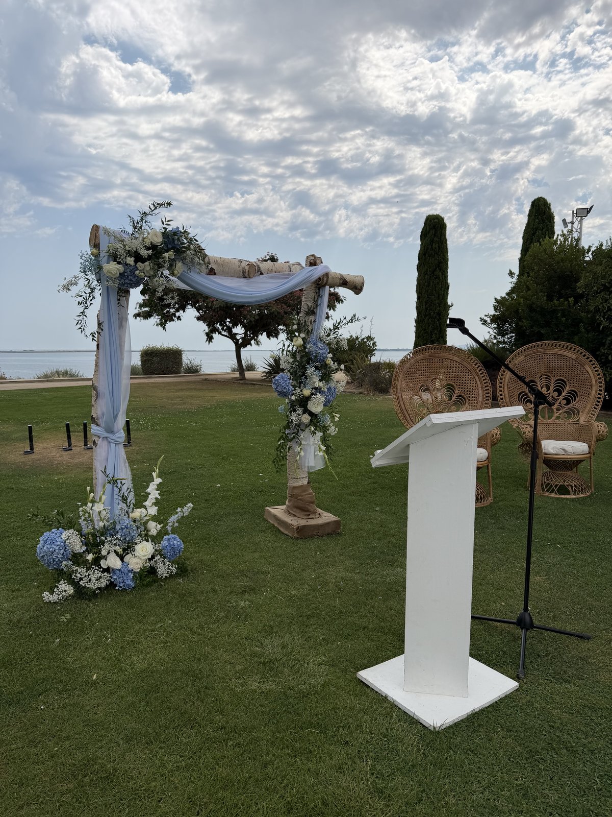 Arche en bouleau drapée de voile bleu clair et fleurie d’hortensias, deux fauteuils Emmanuelle en rotin et pupitre blanc