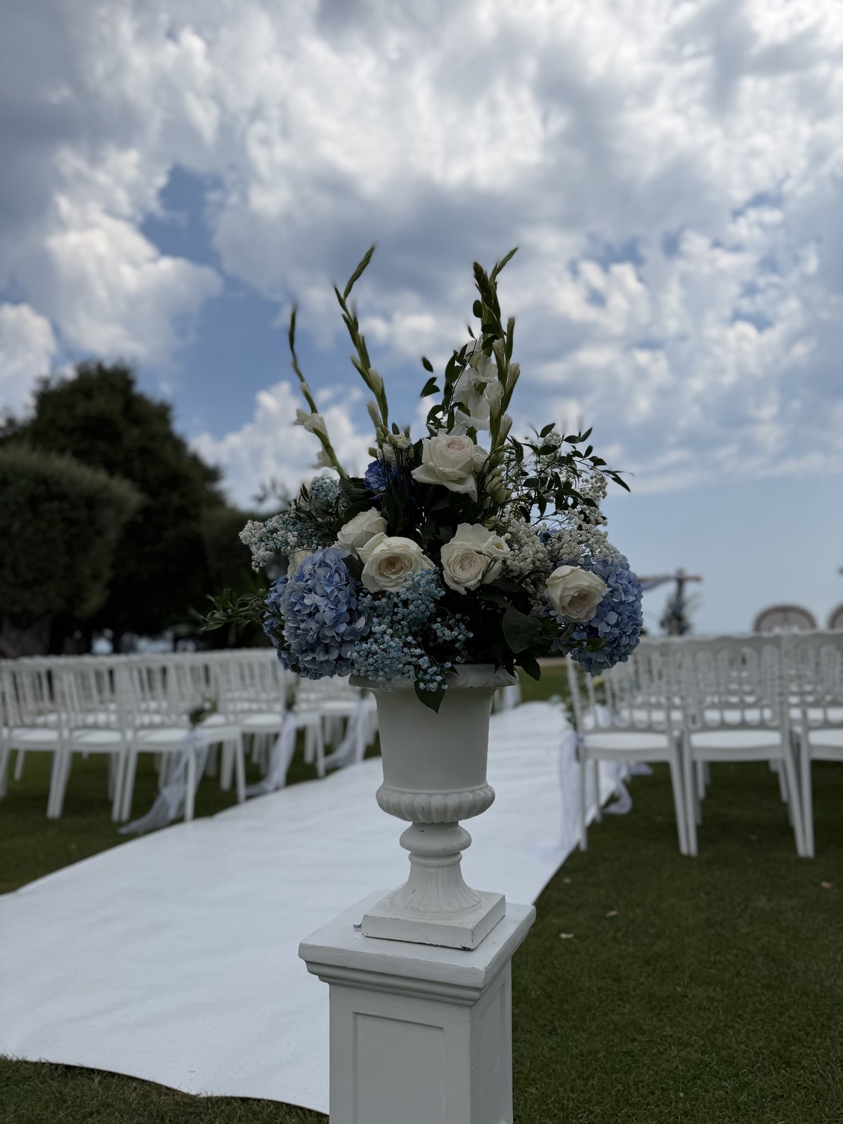 Vase Médicis blanc sur colonne garni d’hortensias bleus, de roses blanches et de glaïeuls, au bord d’une allée blanche