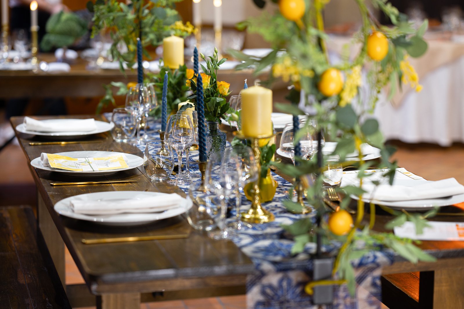 Guirlande de citrons et de feuillage au bout d’une table en bois, assiettes blanches, bougies jaunes et chemin bleu et blanc