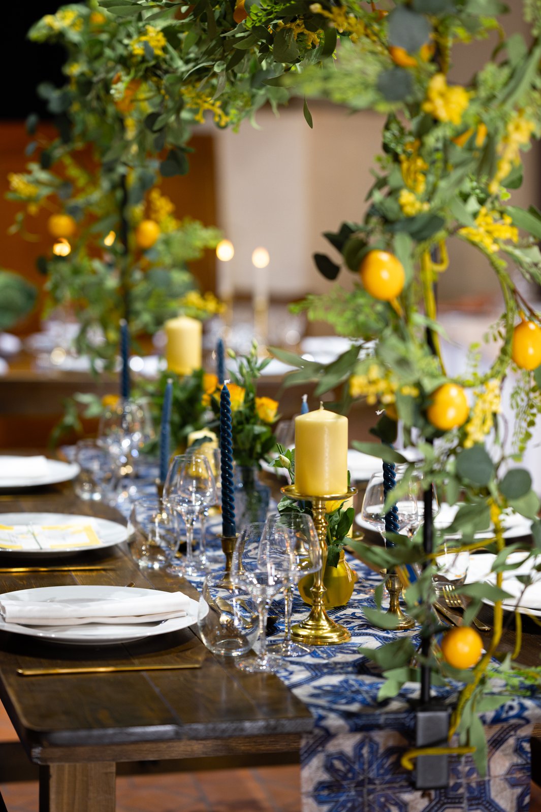 Table en bois vue entre deux montants fleuris de mimosa et de citrons, bougies jaunes sur bougeoirs dorés et verres à pied