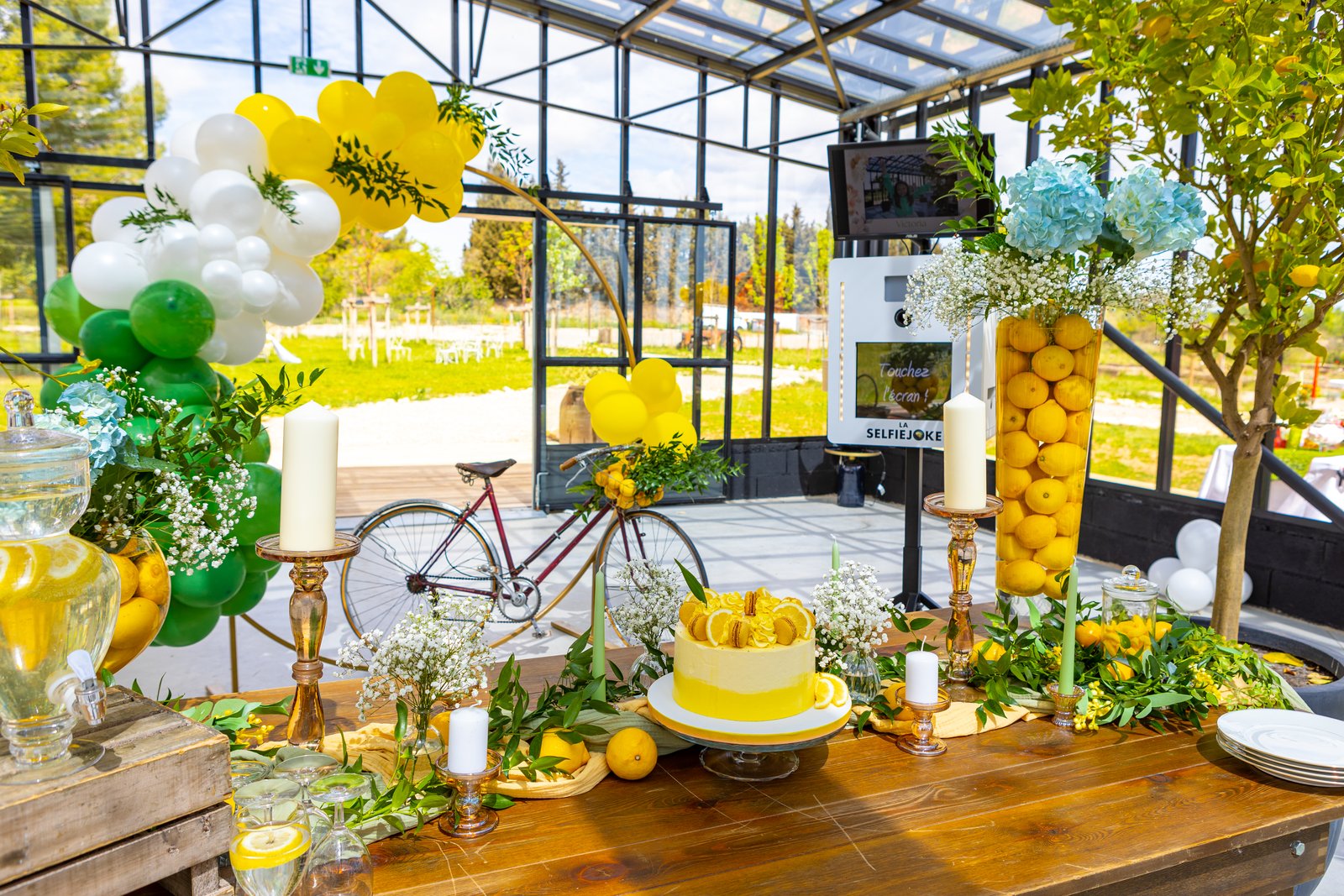 Table en bois sous une verrière, gâteau au citron, bougeoirs ambrés, vase de citrons, vélo ancien et ballons verts et jaunes