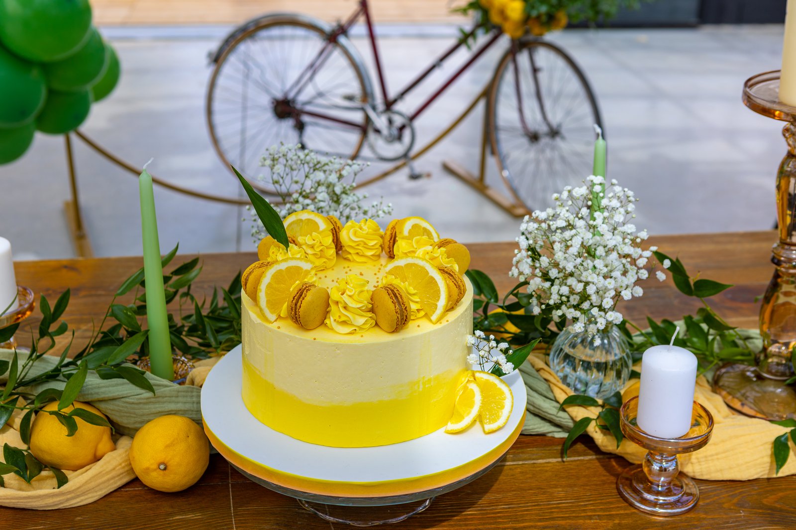 Gâteau jaune aux citrons et macarons sur une table en bois, bougies vertes, gypsophile et vélo ancien en arrière-plan