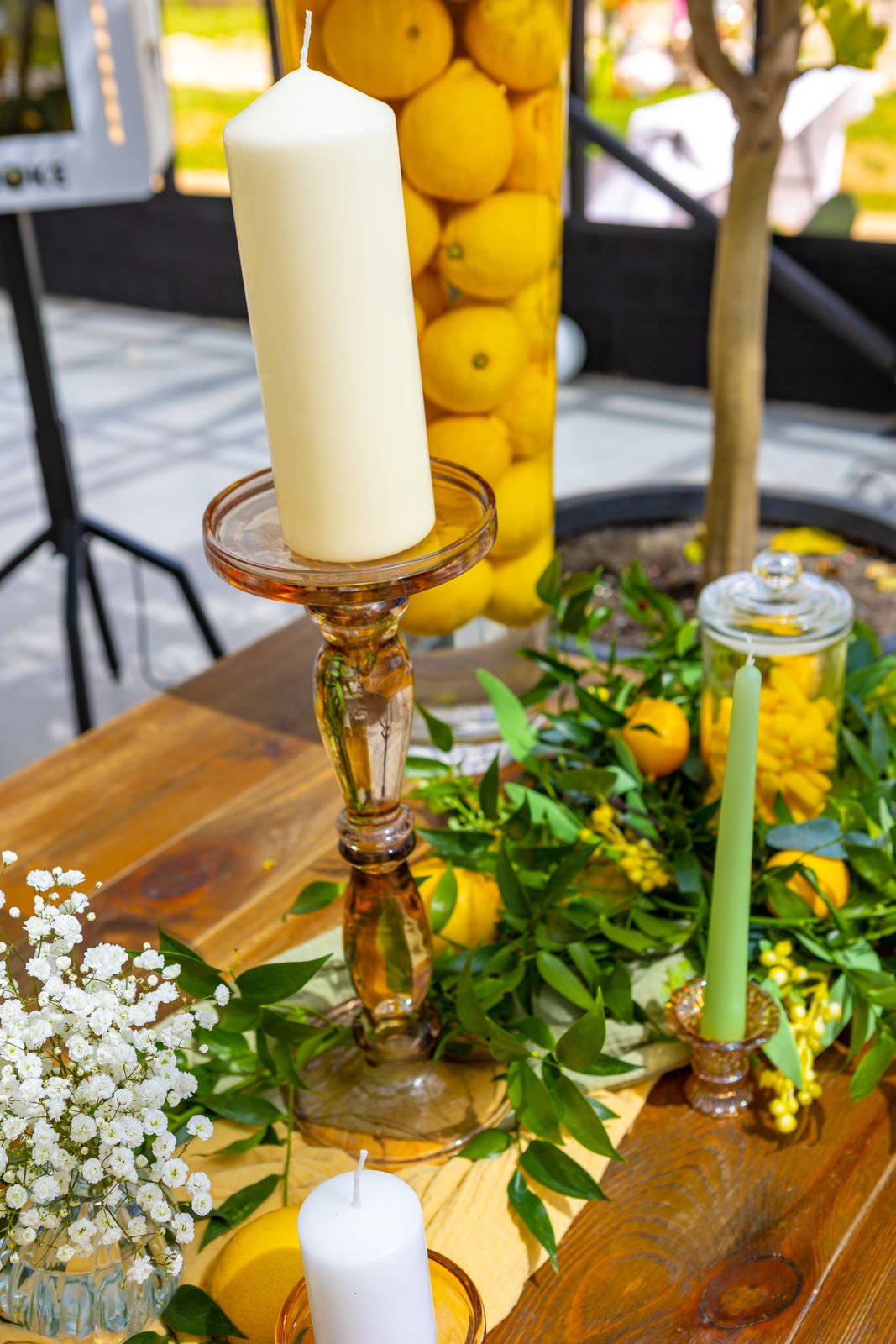 Bougie blanche sur un grand bougeoir en verre ambré, haut vase rempli de citrons, feuillage et bonbonnière sur table en bois