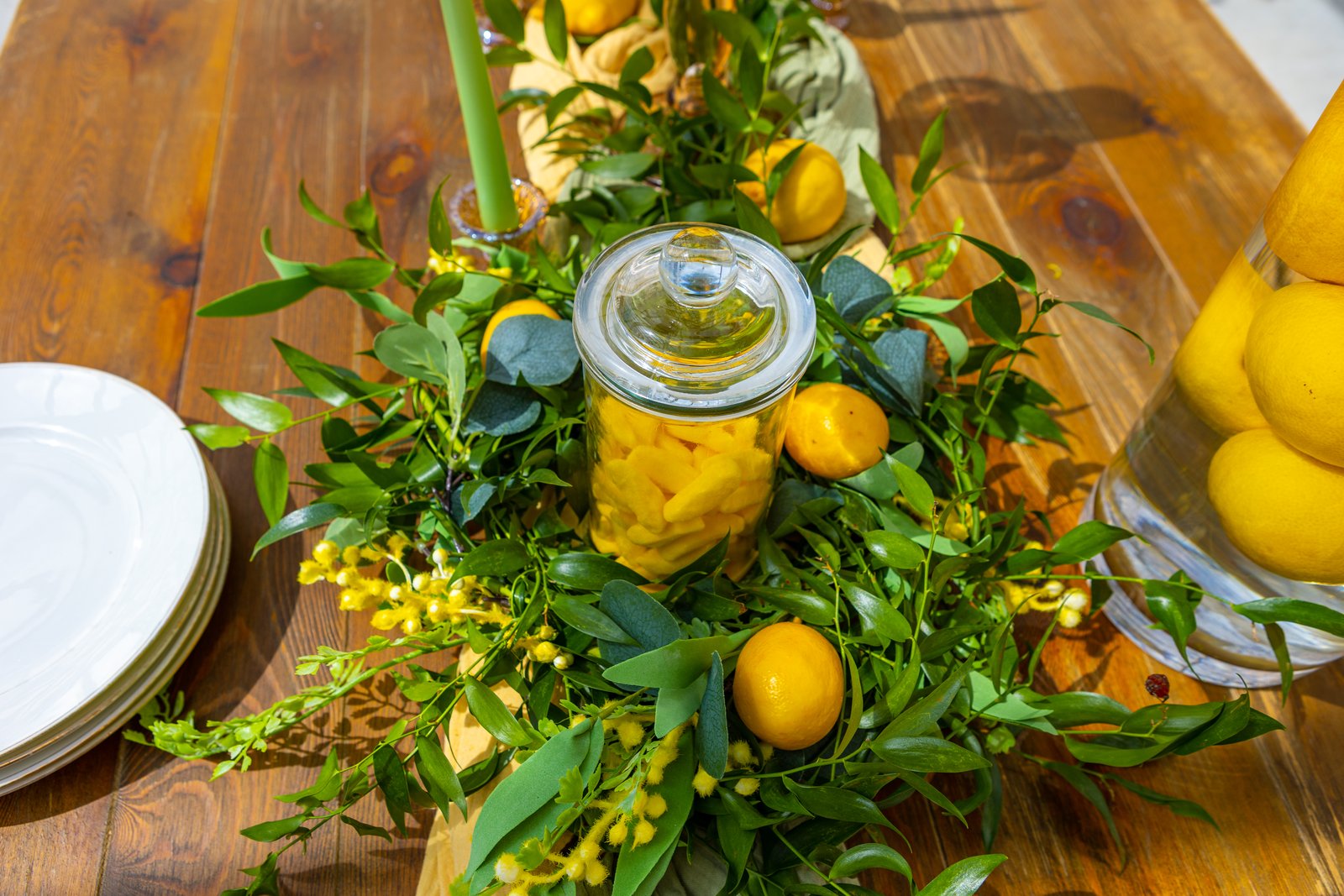 Bonbonnière en verre au milieu d’une couronne de feuillage, de mimosa et de citrons sur une table en bois, assiettes blanches
