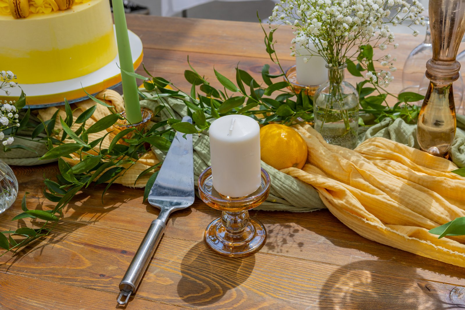 Bougie blanche sur bougeoir en verre ambré, chemins de gaze jaune et vert, citron, feuillage et pelle à gâteau sur table en bois