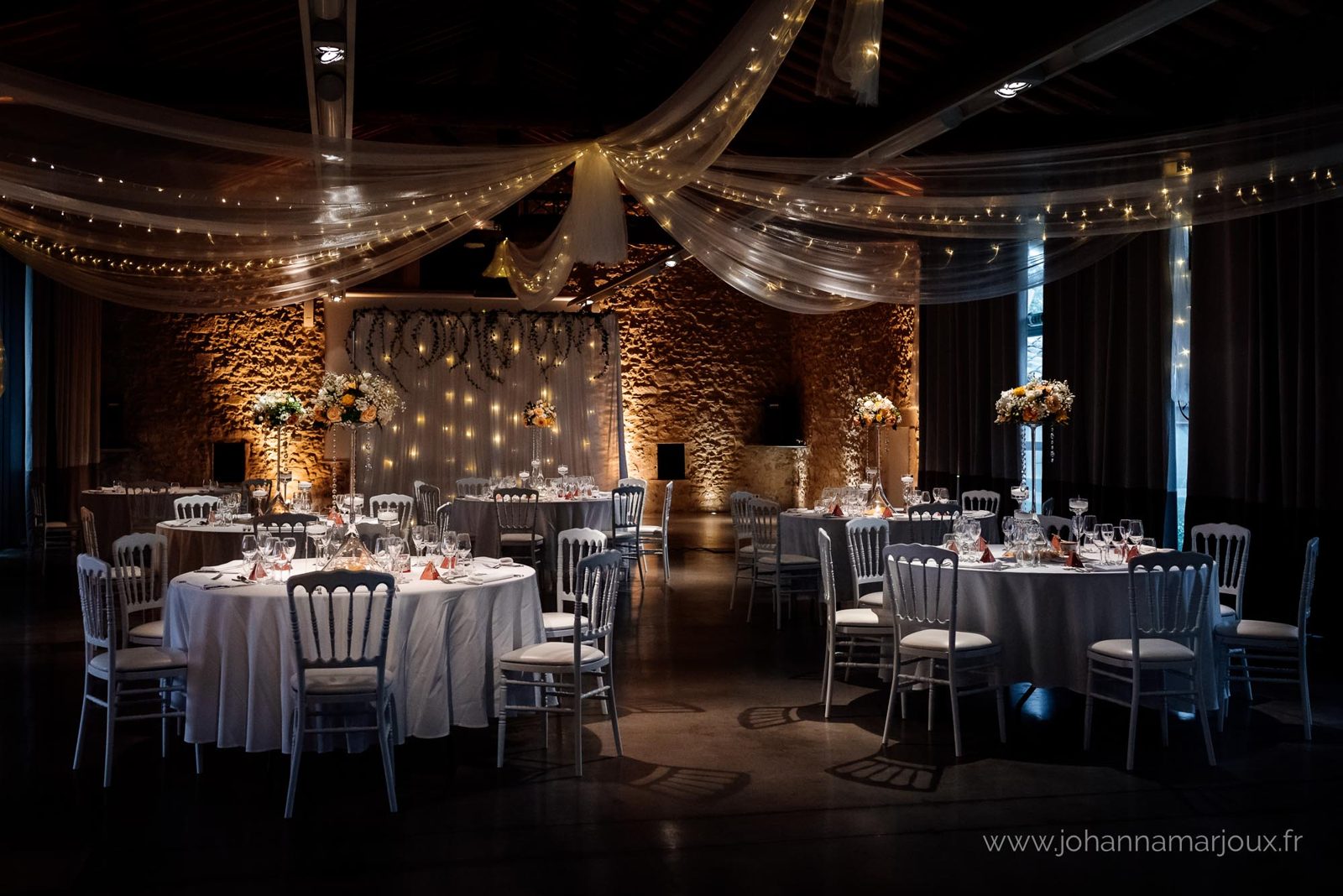 Salle en pierre de nuit, plafond drapé de voilages et guirlandes lumineuses, tables rondes et chaises Napoléon III blanches