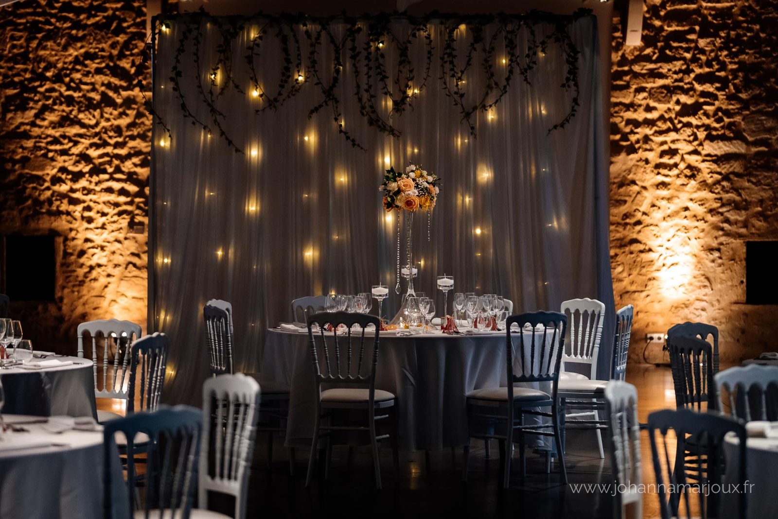 Table ronde devant un rideau lumineux orné de lierre, haute composition florale sur vase en verre, murs en pierre éclairés