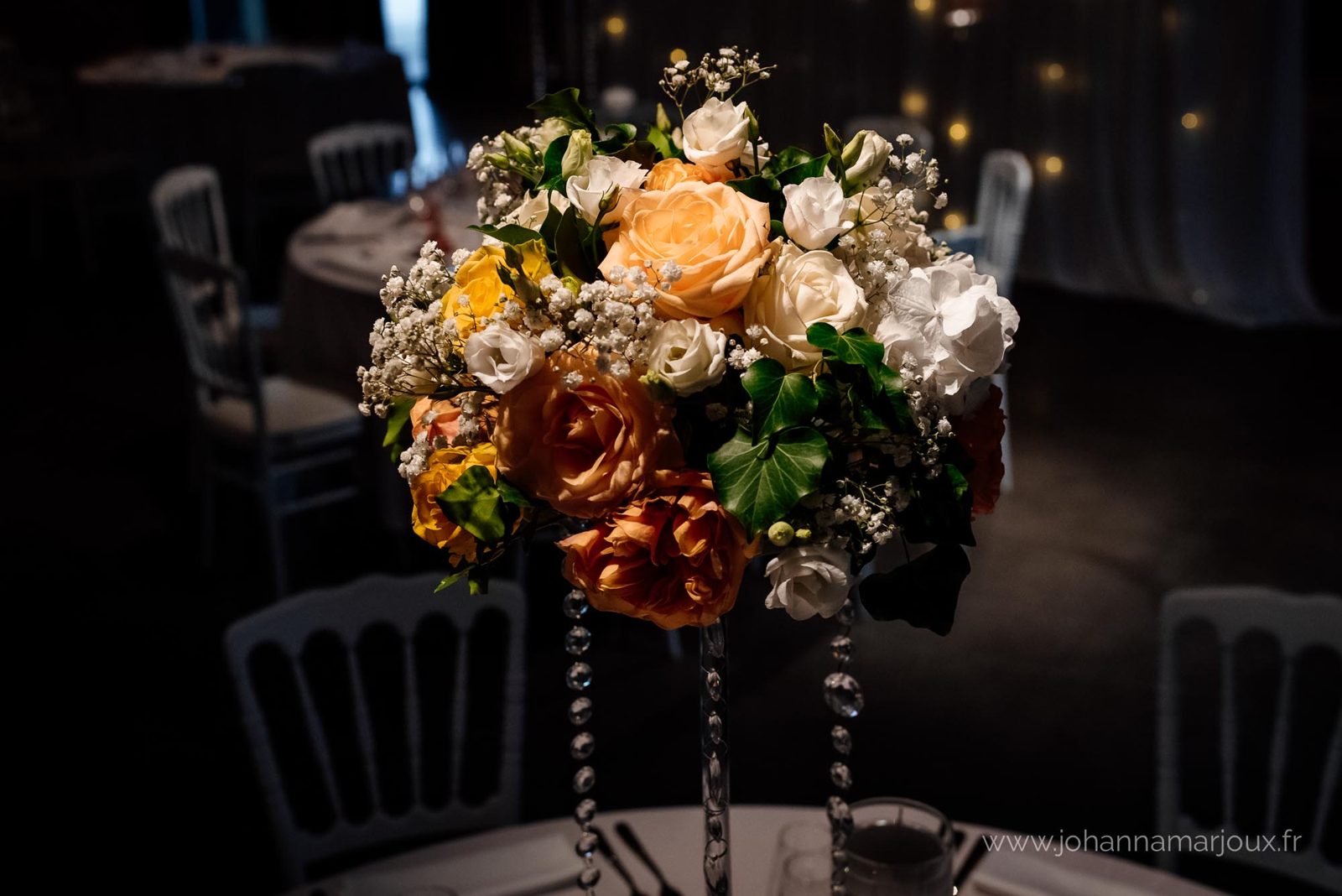 Bouquet de roses pêche, blanches et gypsophile sur un haut vase en verre à pampilles de cristal, chaises blanches autour