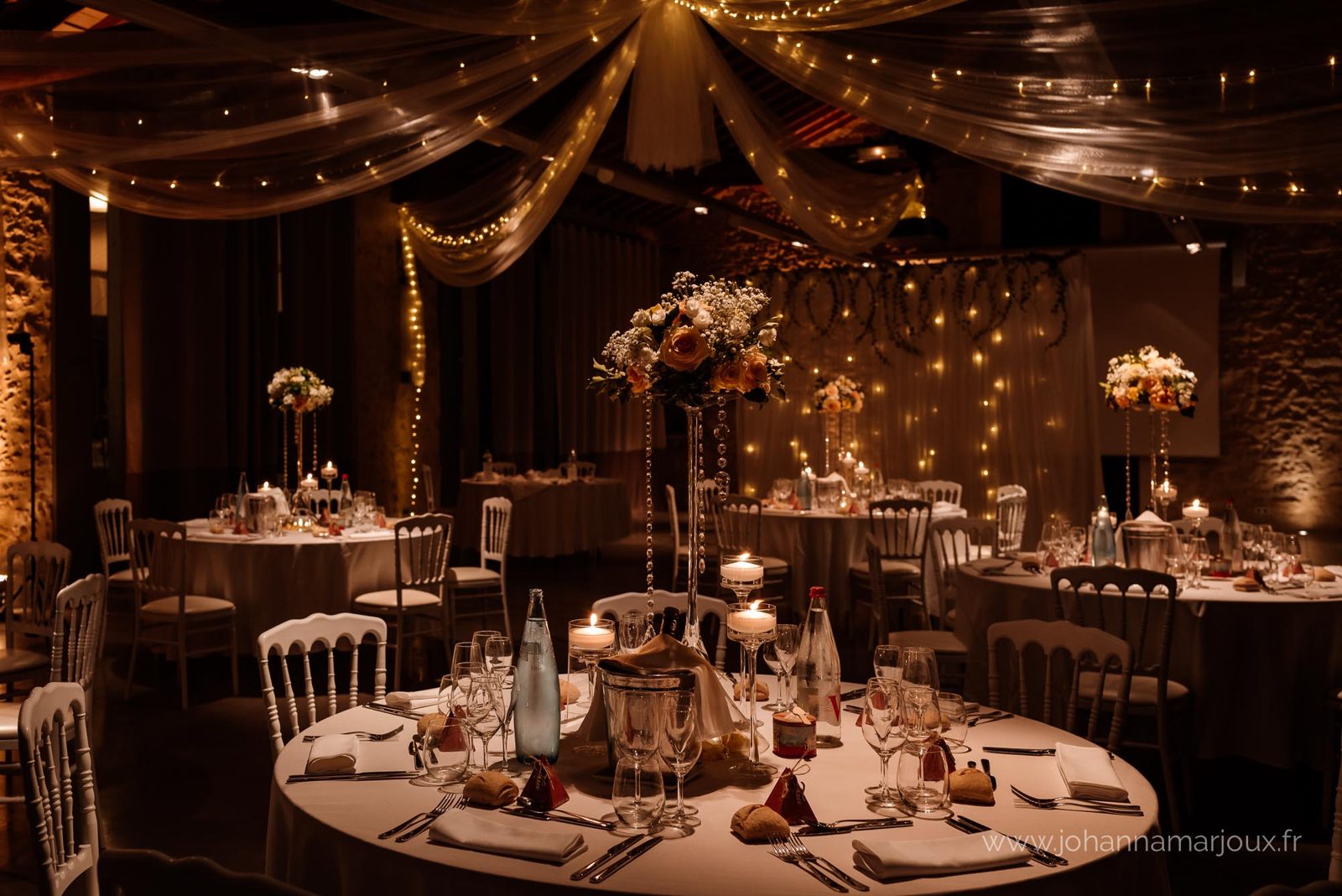 Table ronde dressée avec haute composition florale, bougies flottantes en verres à pied, voilages et guirlandes au plafond