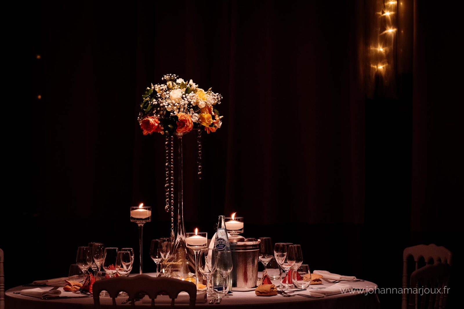 Centre de table de roses orangées sur haut vase en verre, bougies flottantes sur pied et verrerie dans la pénombre