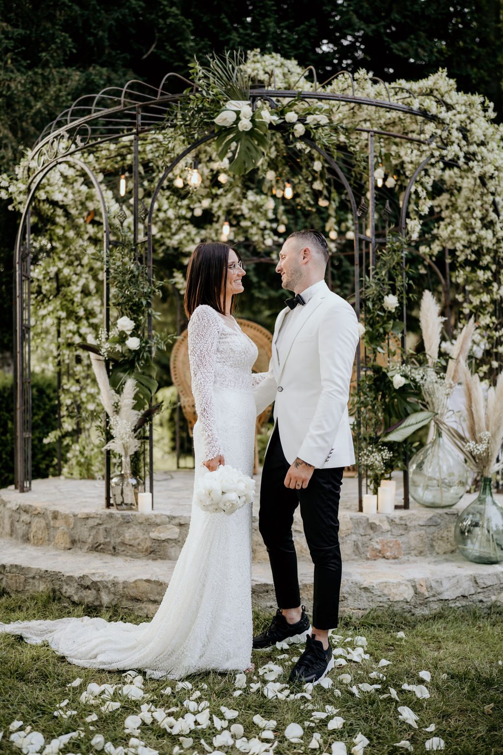 Les mariés sous une gloriette en fer fleurie de blanc, vases dame-jeanne garnis de pampas et allée de pétales