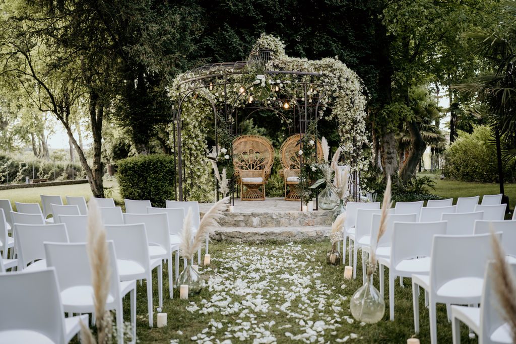 Cérémonie en plein air : gloriette fleurie, deux fauteuils Emmanuelle en rotin, chaises blanches et vases à pampas