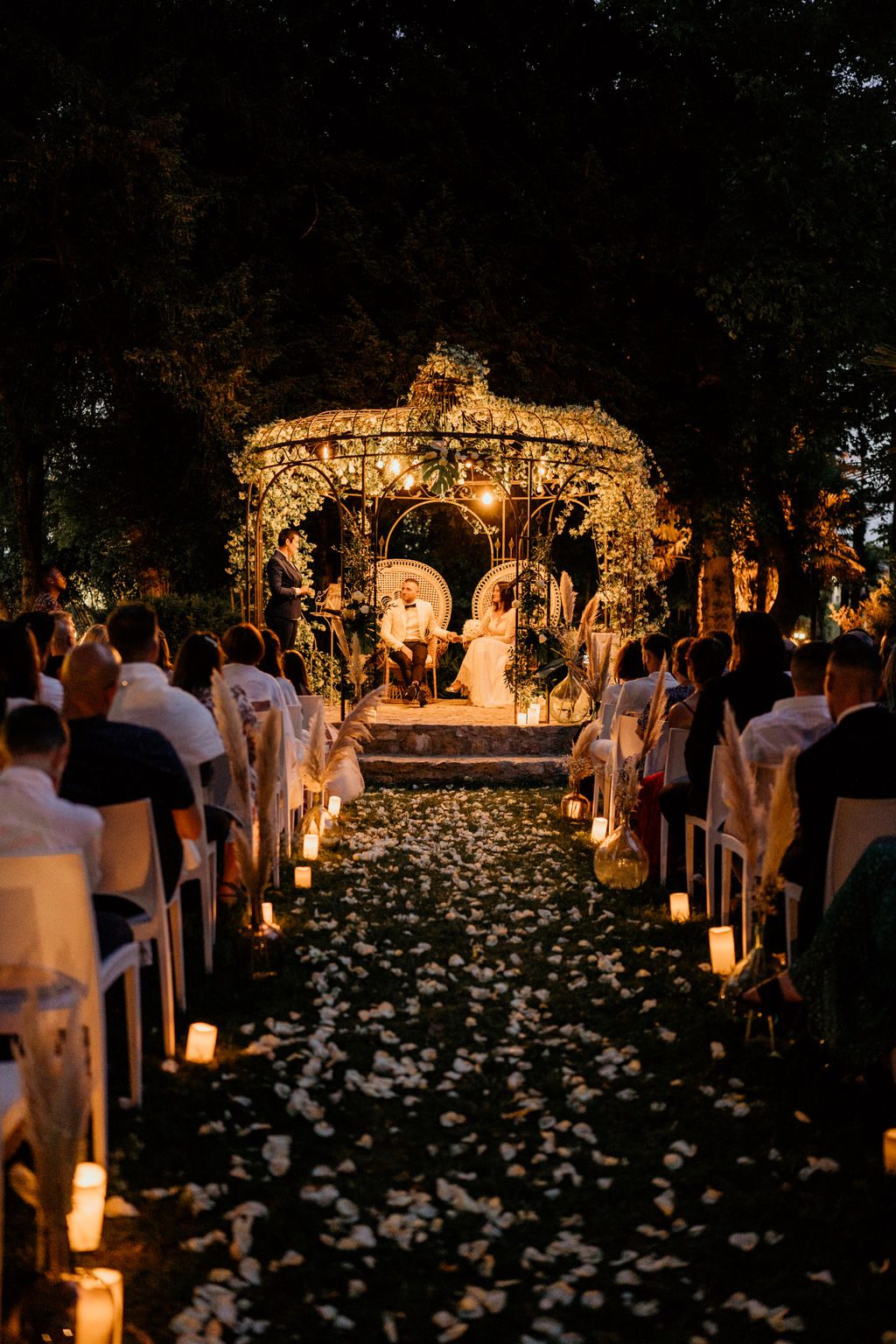 Cérémonie de nuit sous une gloriette illuminée, allée de pétales bordée de bougies et de vases garnis de pampas