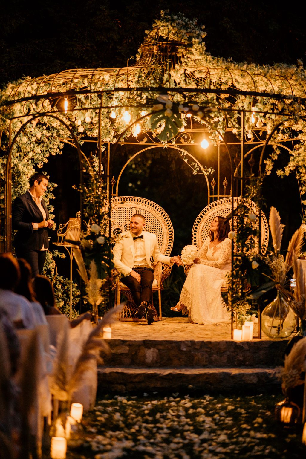 Les mariés assis dans des fauteuils Emmanuelle en rotin sous une gloriette fleurie éclairée d’ampoules, de nuit