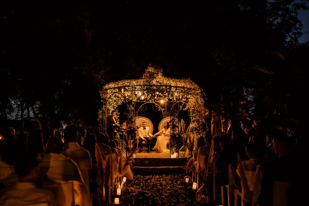Gloriette fleurie illuminée dans la nuit, fauteuils en rotin et allée bordée de bougies entre les rangées d’invités