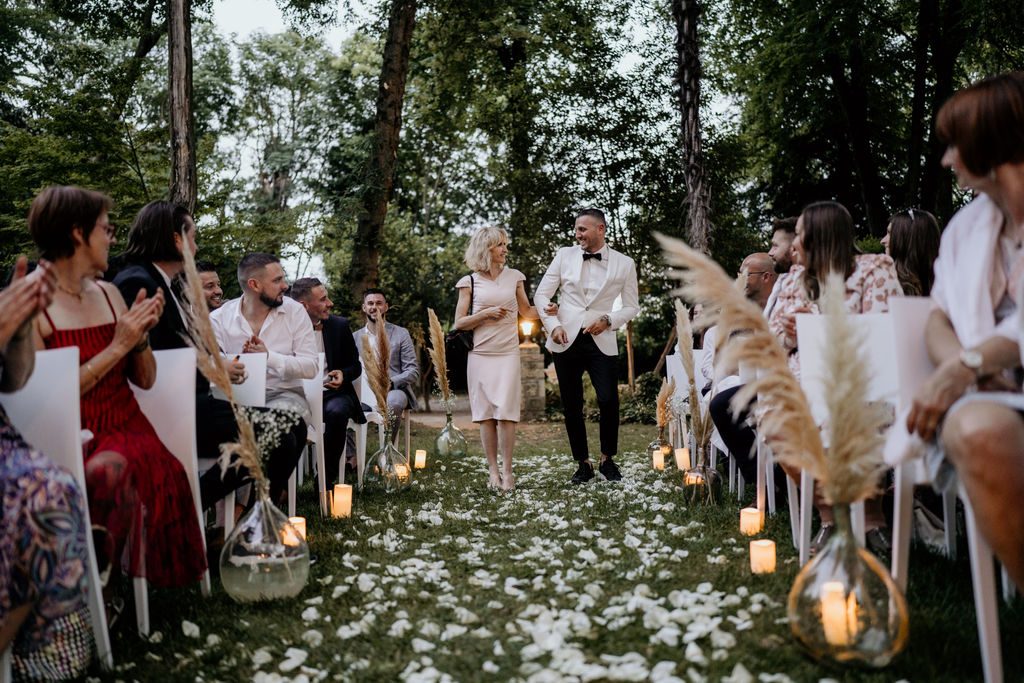 Allée de cérémonie jonchée de pétales blancs, bordée de vases dame-jeanne garnis de pampas et de bougies, invités assis