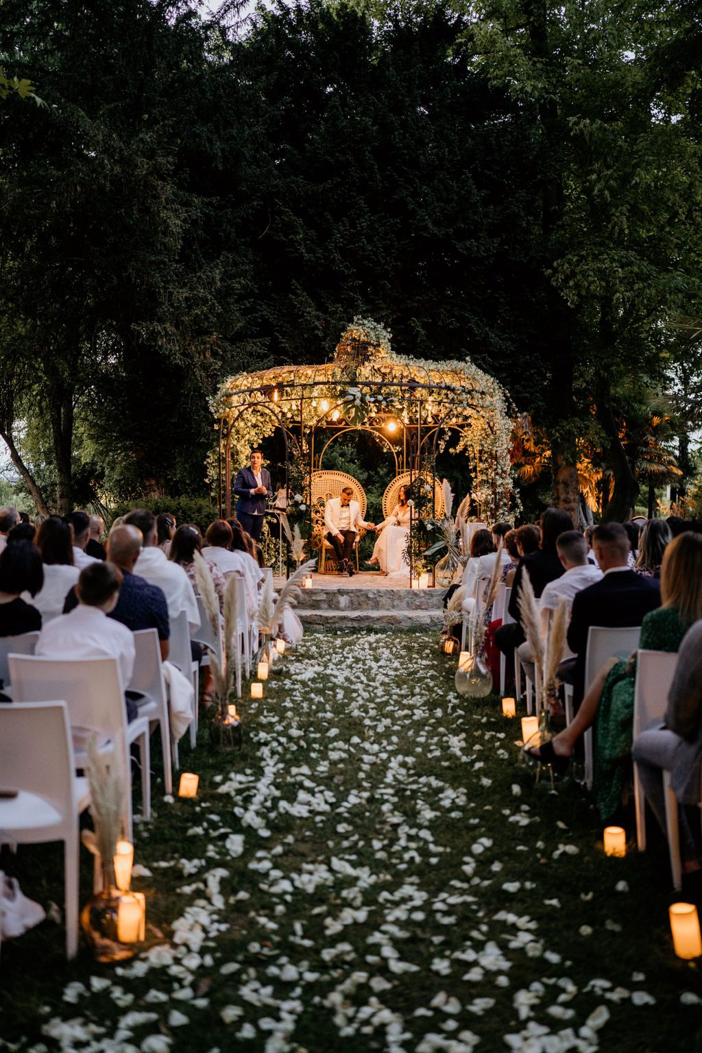 Allée de pétales blancs éclairée de bougies menant à la gloriette fleurie illuminée, chaises blanches de part et d’autre
