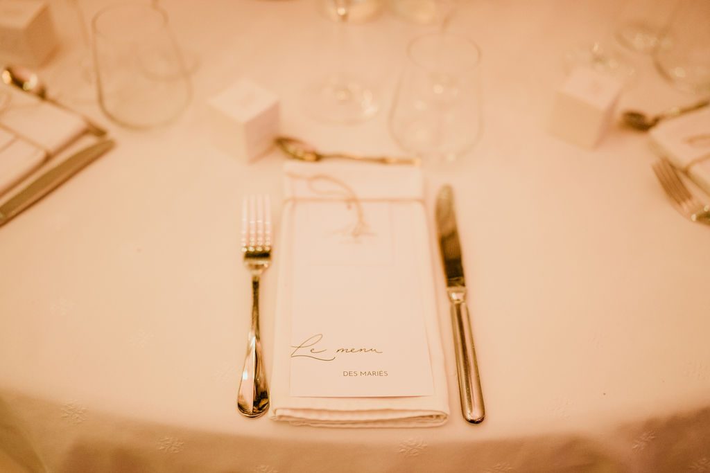 Assiette dressée avec serviette blanche, menu calligraphié noué de ficelle, couverts argentés et verres sur nappe blanche
