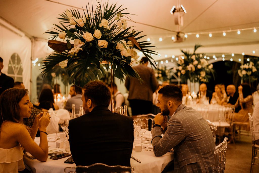 Dîner sous chapiteau éclairé de guirlandes guinguette, centres de table de palmes et roses blanches en hauteur