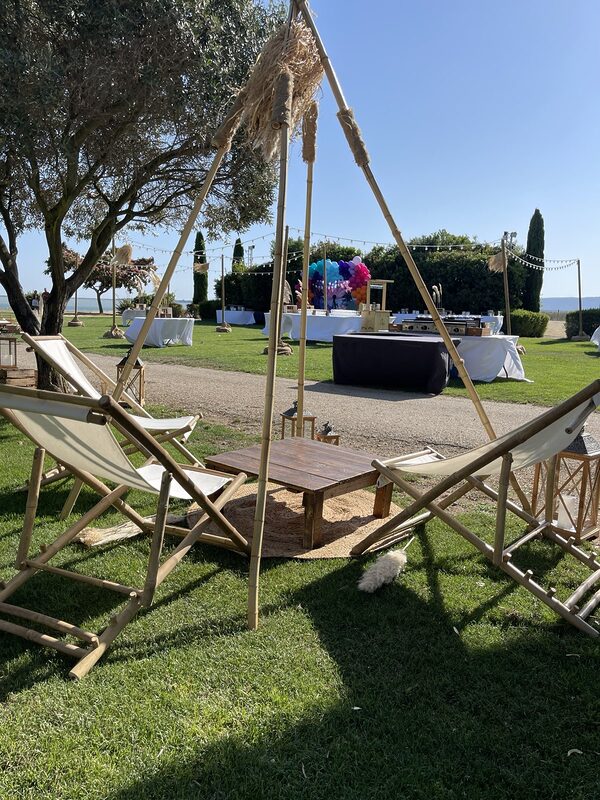 Chiliennes en bambou autour d’une table basse en bois sur un tapis rond en jute, sous un tipi en bambou orné de pampas, lanternes en bois