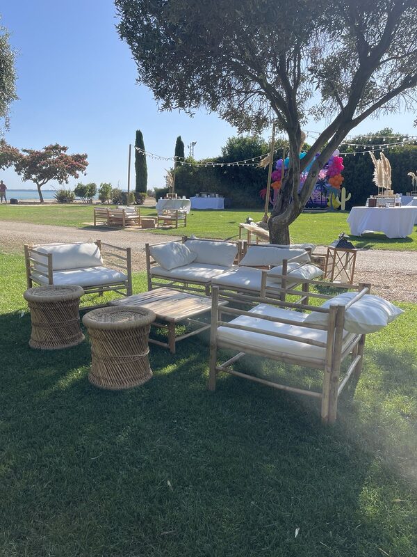 Salon en bambou aux coussins écrus et poufs en bambou tressé à l’ombre d’un olivier, guirlandes guinguette sur mâts en bois au fond