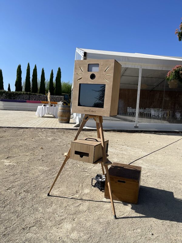 Borne photo en bois sur trépied devant un chapiteau, tonneau en bois et table nappée de blanc en arrière-plan