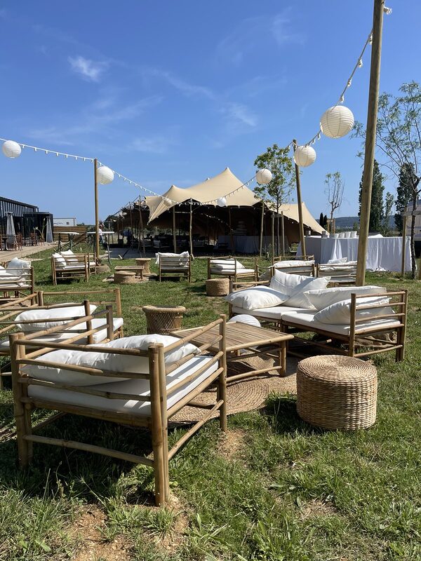 Salons en bambou aux coussins blancs, poufs tressés et tapis de jute sur la pelouse, guirlandes guinguette et lampions blancs sur mâts en bois, tente tendue au fond
