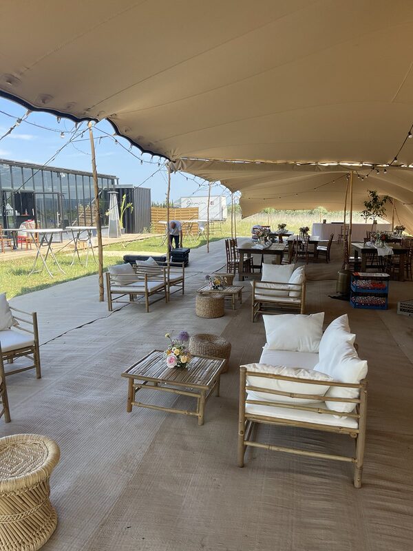 Sous une tente tendue : salons en bambou, tables basses fleuries et poufs tressés, longues tables en bois au fond, guirlandes guinguette