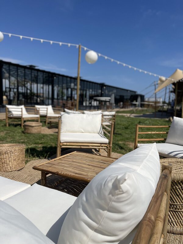 Gros plan sur un canapé en bambou à coussins blancs et une table basse, lampions blancs et guirlande guinguette dans le ciel bleu