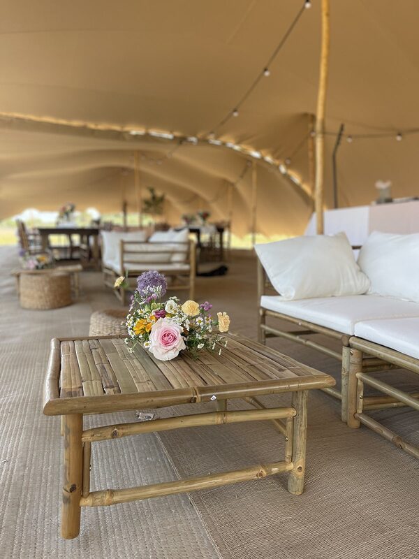 Table basse en bambou garnie d’un bouquet champêtre, canapé blanc et poufs tressés sous la tente