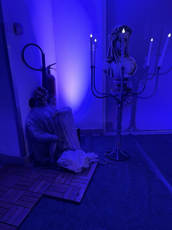 Spectre squelette assis au pied d’un mur et chandelier argenté à cinq bougies LED autour d’un buste voilé, sous lumière bleue