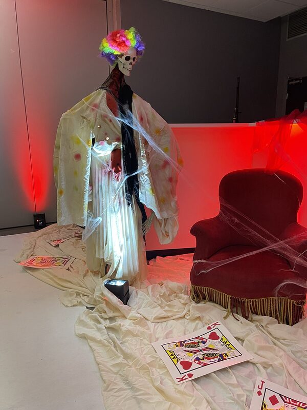 Clown squelette à perruque arc-en-ciel et fauteuil en velours rouge à franges, cartes à jouer géantes au sol, lumière rouge