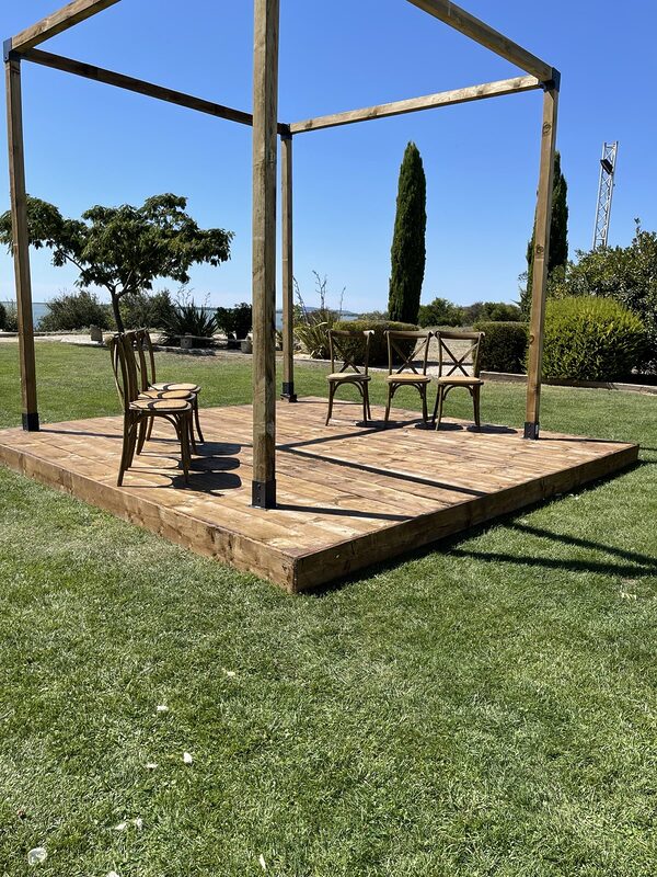 Scène en bois brut et houppa carrée vues de biais, chaises Crossback à assise cannée, cyprès et étang
