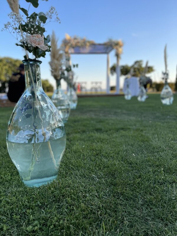 Gros plan sur un vase en verre au pied de l’allée, pampas et rose pâle, houppa fleurie floue au fond