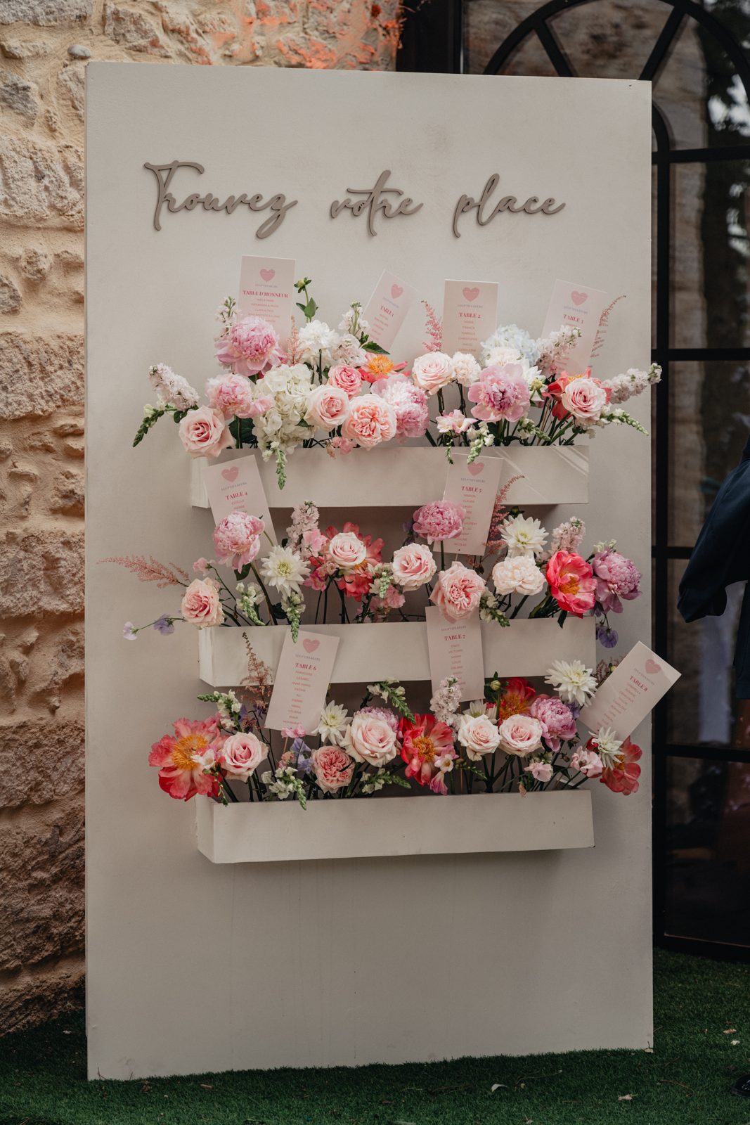 Plan de table « Trouvez votre place » : panneau blanc garni de jardinières de roses et pivoines roses, cartes de table