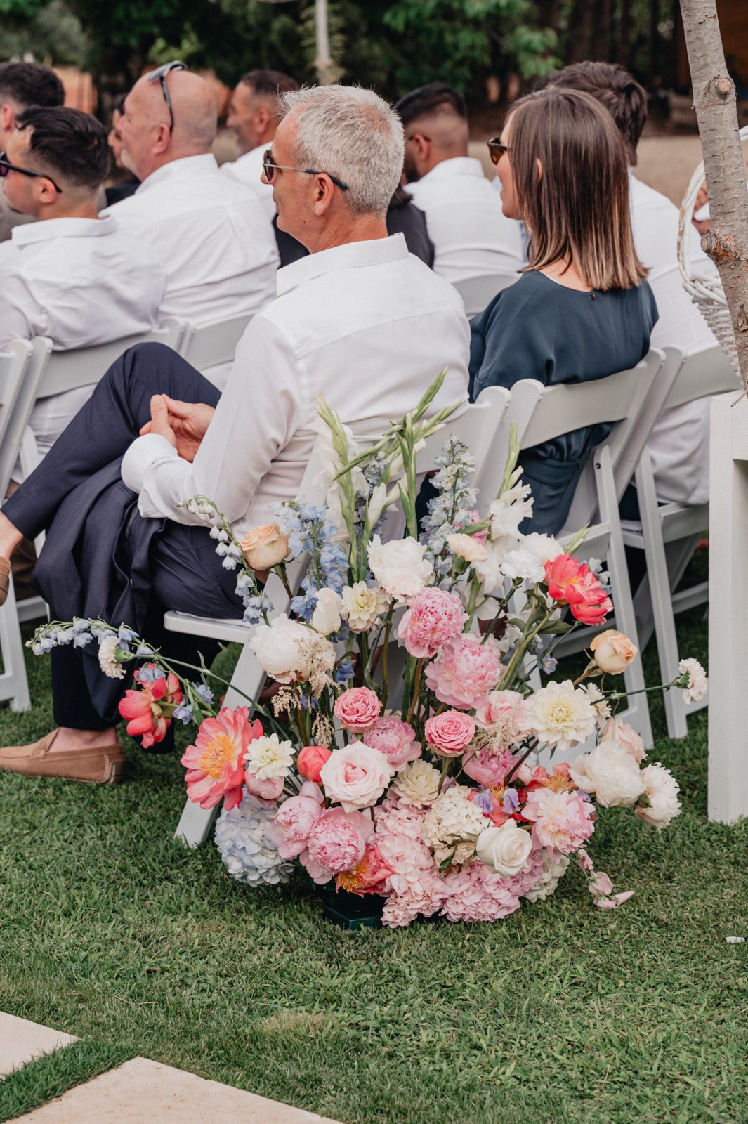 Bouquet de sol de pivoines, roses et fleurs pastel au bout d’une rangée de chaises blanches, invités assis au jardin