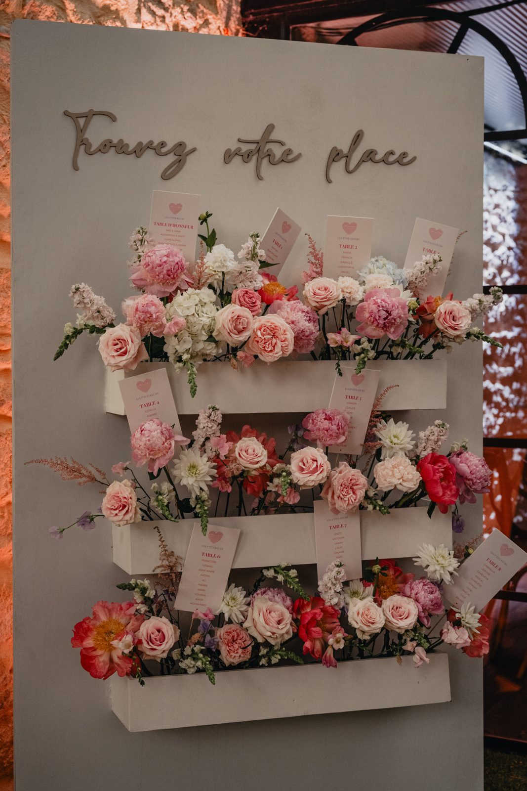 Panneau blanc « Trouvez votre place » avec trois jardinières de roses et pivoines, cartes de table roses sous une verrière