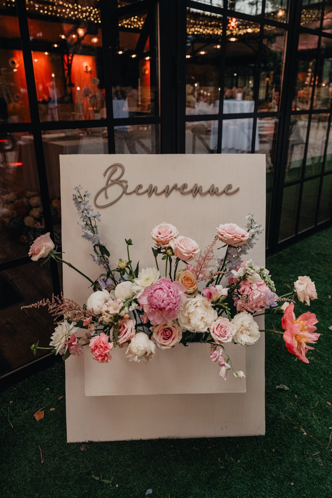 Panneau de bienvenue blanc lettré « Bienvenue », garni d’une jardinière de pivoines et roses, posé sur le gazon
