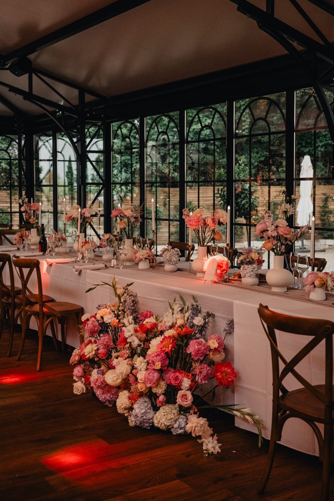 Table d’honneur nappée de blanc sous verrière, vases blancs fleuris, grandes bougies et bouquet de sol de pivoines