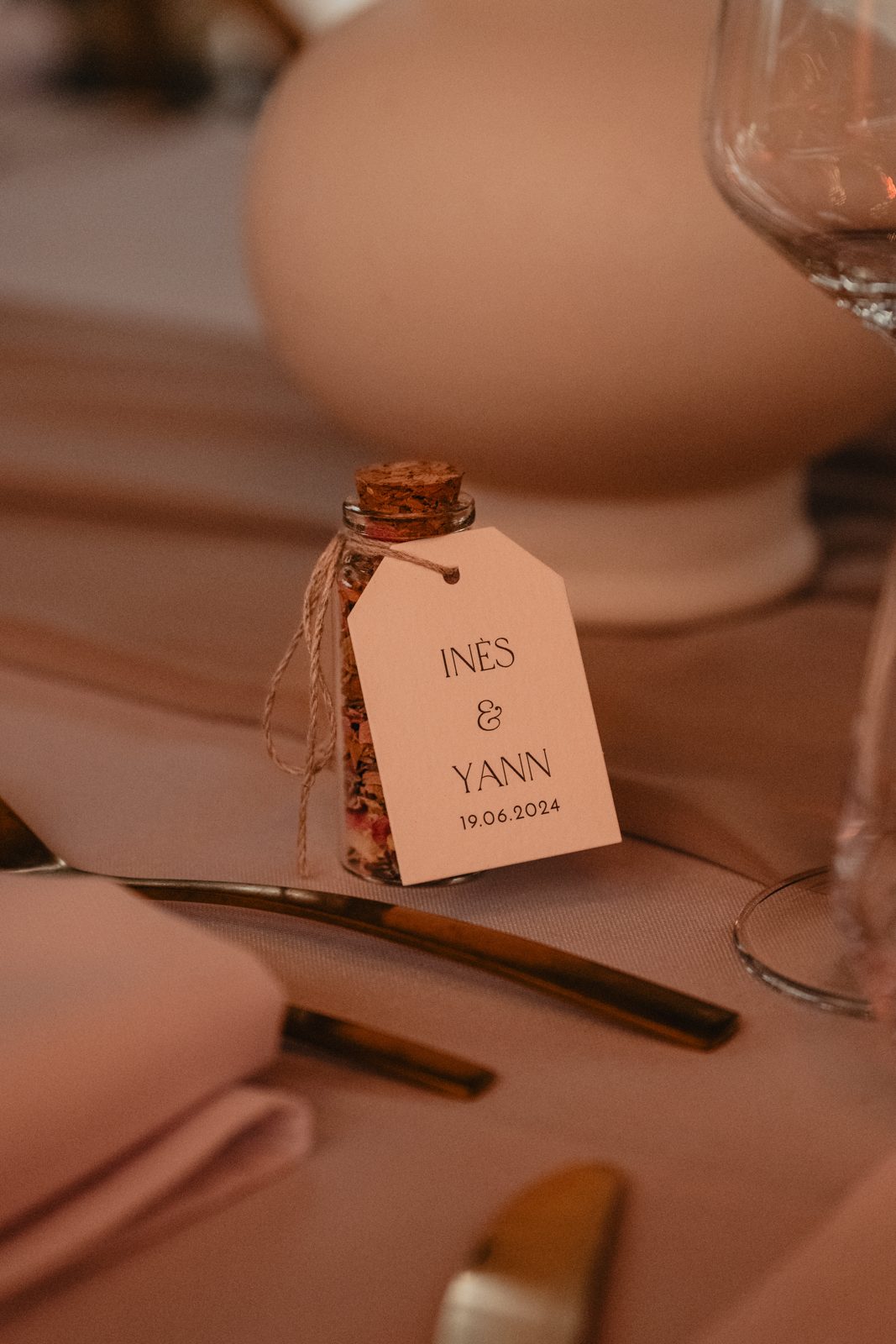 Cadeau d’invité : petite fiole de pétales séchés avec étiquette kraft sur une table nappée de rose poudré, couverts dorés